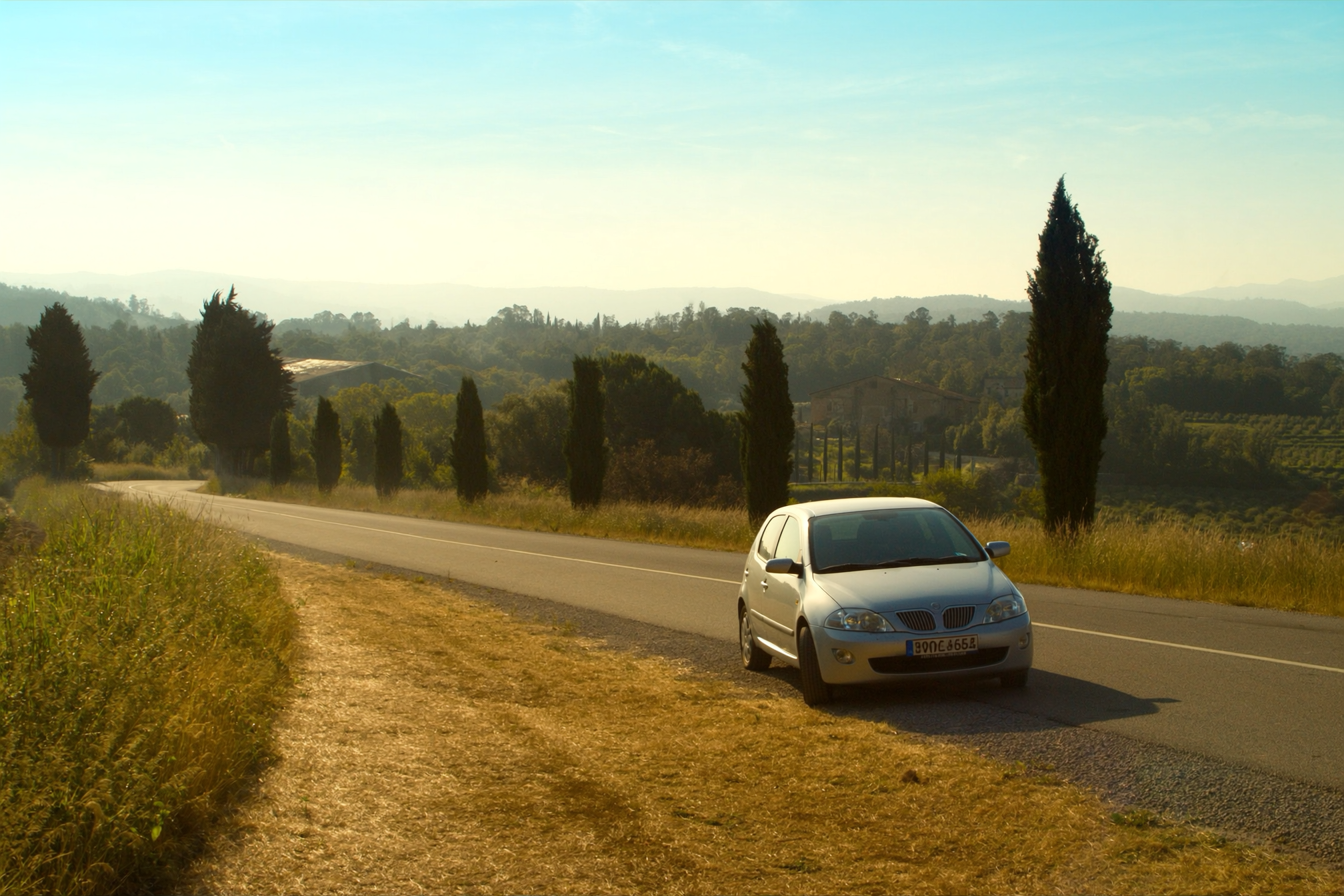 A rental car parked on the gravel shoulder of a cypress lined road in Val d'Orcia, Tuscany, at golden hour