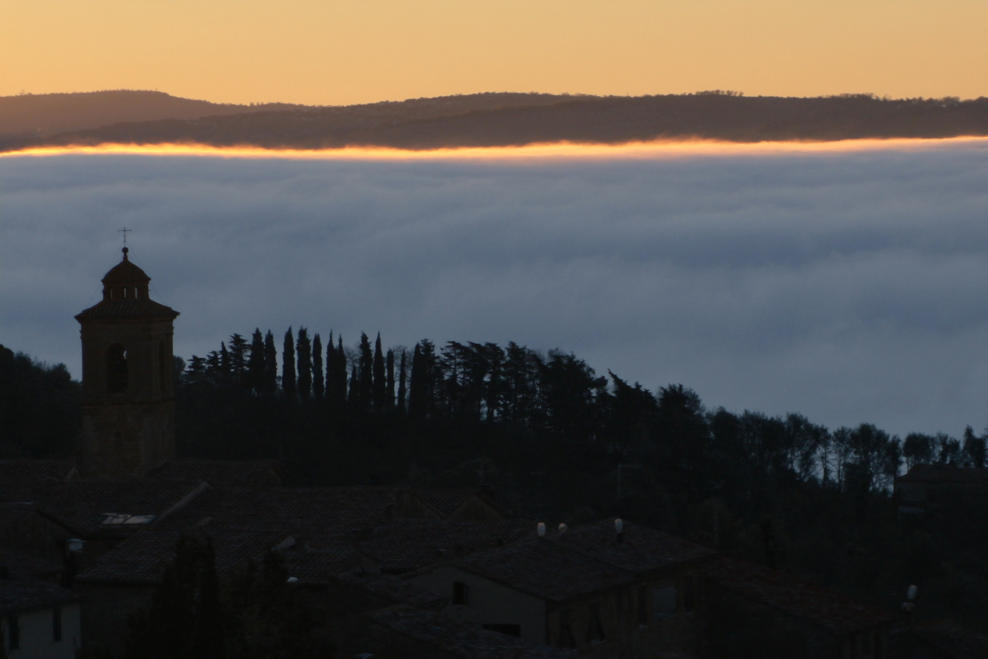 Autumn fog pooling in the Val d'Orcia valley below a hilltop town at sunrise, with cypress trees rising through the mist